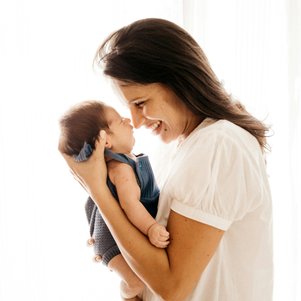 A woman holding a baby and smiling at it, creating a tender moment. Both are dressed in light-colored clothing with natural light in the background.